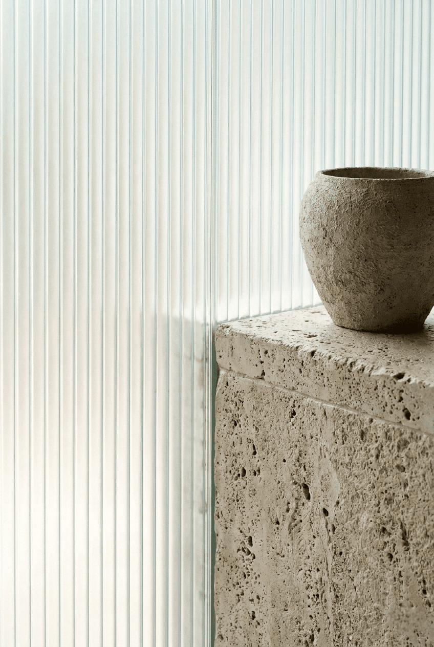 Raw wabi-sabi ceramic vase resting on a massive unpolished limestone ledge against a frosted reeded glass wall